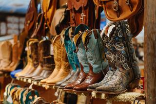 A colorful array of cowboy boots lined up, showcasing various styles and intricate designs, with leather accessories in the background.