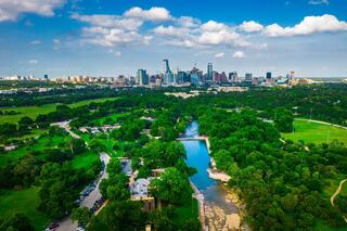 A vibrant city skyline rises above lush green parks and a winding river, set against a backdrop of blue sky and scattered clouds.