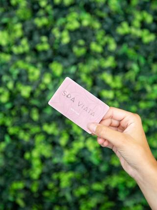 A hand holds a pink card with "SPA VIATA" printed on it, against a vibrant green foliage background.