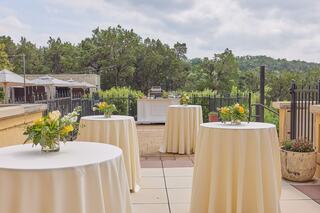 A serene outdoor setting with several elegantly arranged tables topped with floral centerpieces, surrounded by lush greenery and distant hills.