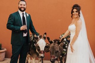 A smiling couple in formal attire stands beside two adorned donkeys, celebrating with joy against a warm-colored backdrop.