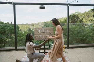 A man kneels with a ring in hand, proposing to a woman in a flowing dress, surrounded by greenery and a "Will you marry me?" sign.