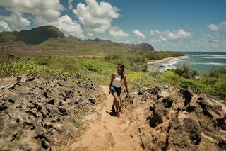 A woman hikes along a sandy path bordered by rocky terrain, with lush greenery and a stunning coastal view under a bright blue sky.