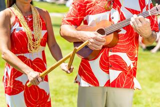 A woman in a floral dress holds wooden sticks while a man in a matching shirt strums a ukulele, set in a vibrant outdoor setting.