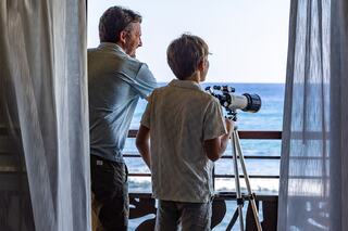 A man and a boy look through a telescope from a balcony, gazing at the ocean on a sunny day. Curtains frame their view.