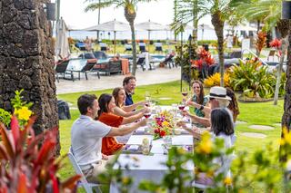 A group of friends celebrates at an outdoor table, raising glasses in a lush, tropical setting, surrounded by vibrant flowers and greenery.