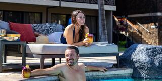 A woman lounges by the pool with a drink, while a man relaxes in the water, enjoying a sunny day together. Bright, tropical vibe.