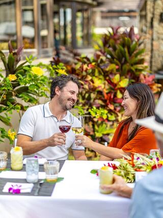 A couple joyfully toasts with drinks at a beautifully set table amidst vibrant greenery and colorful flowers, enjoying each other's company.