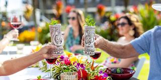 Two people toast with ornate tiki mugs at a vibrant outdoor gathering, surrounded by friends, colorful flowers, and delicious food.