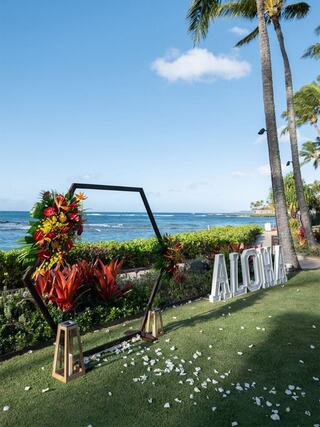 A tropical beach setting features a floral arch, greenery, and the word "ALOHA" in large letters, creating a vibrant, inviting atmosphere.