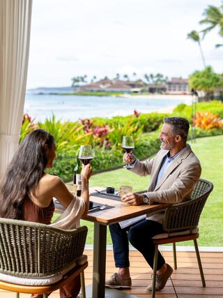 A couple enjoys a toast at a scenic restaurant, overlooking lush gardens and a calm sea, creating a relaxed and romantic atmosphere.