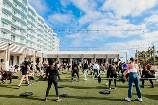 A large group participates in an outdoor fitness class, using weights on a grassy area, against a backdrop of modern architecture and clear skies.