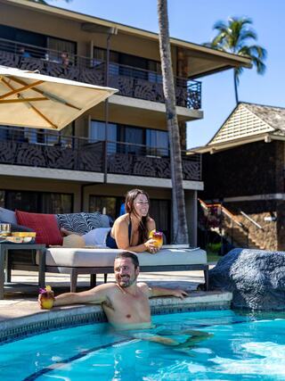 A relaxed duo enjoys tropical drinks by a pool, soaking up the sun with palm trees and a cozy resort backdrop.