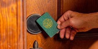 A hand holds a key card with a tree logo, poised to unlock a wooden door with a round keyhole.