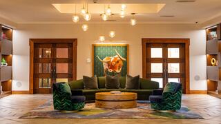 A cozy lobby features a green sofa with patterned chairs, a round wooden table, and a vibrant bull painting, illuminated by stylish pendant lights.