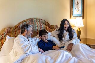 A laughing couple in cozy robes enjoys a playful moment with a boy, all relaxing on a bed while watching TV together.