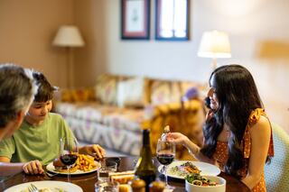 A woman shares a meal and smiles with a young boy at a dining table, while another person sits off to the side in a cozy, homey setting.