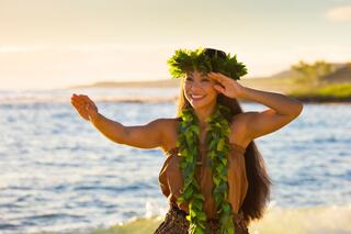 A woman gracefully dances by the shore, adorned with a flower crown and green lei, radiating joy against a scenic coastal backdrop.