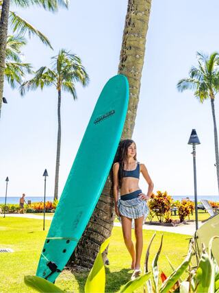 A young woman stands by a palm tree, leaning against a vibrant teal surfboard, with lush greenery and the ocean in the background.