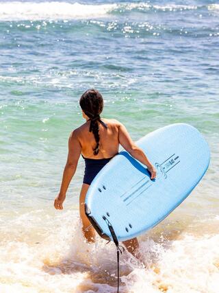 A person in a swimsuit walks into the ocean, carrying a blue surfboard, ready to embrace the waves. Sunlight sparkles on the water.