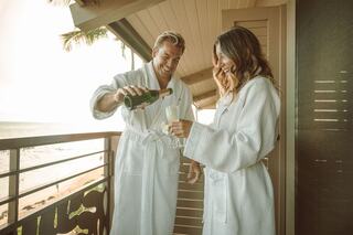 A couple in white robes joyfully pours champagne on a balcony, with a beautiful ocean view in the background. Sunlight enhances the moment.