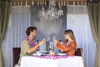 A couple enjoys a romantic dinner, wearing leis and toasting with drinks at an elegantly set table under a chandelier.