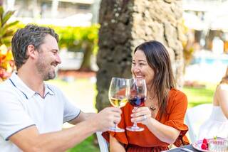 A happy couple clinks glasses, enjoying drinks outdoors in a sunny setting, surrounded by greenery and the ambiance of a lively gathering.