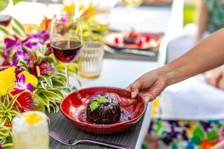 A hand presents a rich chocolate dessert adorned with mint on a vibrant red plate, surrounded by tropical flowers and glasses of wine.