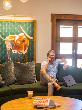 A man in a light blue shirt sits on a green couch, talking on the phone, with a laptop and drinks on a wooden table nearby.