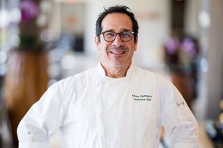 A bald man with a beard stands confidently in a chef's coat, posed against a backdrop of greenery and a white fence.