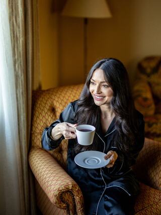 A woman in a black pajama set smiles while holding a cup of tea, seated comfortably in an inviting armchair by a window.