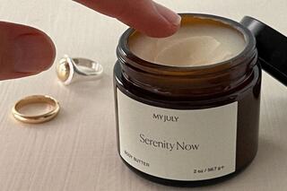 A finger dips into a jar of "Serenity Now" body butter, surrounded by two rings on a soft surface, suggesting a moment of self-care.