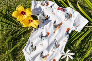 A playful baby onesie features colorful rooster prints, surrounded by vibrant yellow hibiscus flowers on green grass.