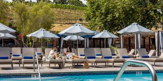 A couple relaxes by a luxurious pool, enjoying drinks under umbrellas with a scenic vineyard in the background on a sunny day.