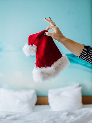A hand holds a festive red Santa hat with white fur trim against a soft blue background, suggesting a cheerful holiday vibe.