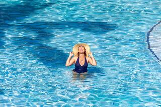 A woman in a dark swimsuit and wide-brimmed hat smiles playfully while standing in a sparkling blue pool. Sunlight dances on the water's surface.