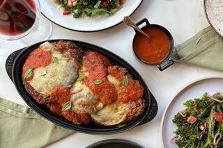A dish of breaded eggplant topped with marinara sauce and melted cheese, garnished with herbs, served alongside a fresh salad and red wine.