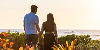 A couple holds hands, standing together on a beach while watching the waves at sunset, surrounded by lush greenery.