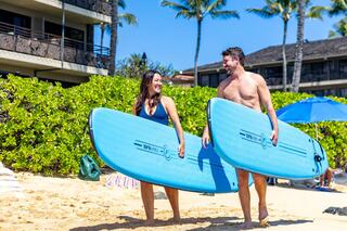 Two cheerful surfers carry blue surfboards along a sunny beach, surrounded by palm trees and beachgoers, ready for a fun day on the waves.