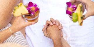 Two hands are clasped together, surrounded by tropical drinks adorned with pineapple and orchids, evoking a sense of connection and relaxation.