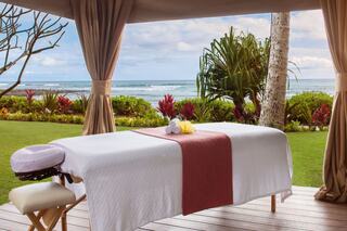 A tranquil outdoor spa setup with a massage table, overlooking a serene ocean view and lush tropical plants in the background.