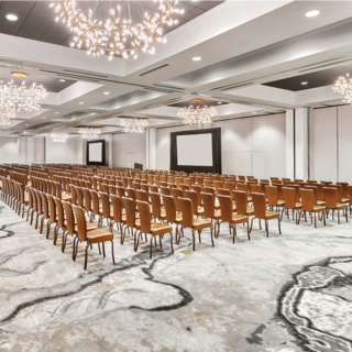 A spacious conference hall featuring rows of brown chairs, elegant light fixtures, and a neutral carpet, suited for events or presentations.