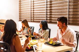 A group enjoys a lively dinner at a well-lit table, sharing food and laughter, with glasses of wine and a warm, inviting atmosphere.