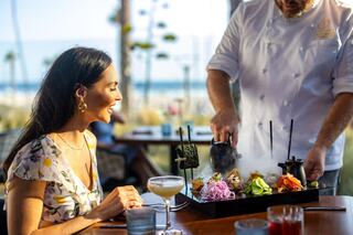 A woman watches in delight as a chef presents a colorful dish, releasing steam, against a backdrop of a sunny beach.