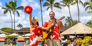 A joyful duo dressed in vibrant Hawaiian attire performs with a guitar, surrounded by palm trees and a tropical resort backdrop.