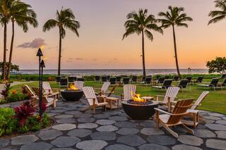 A serene patio with fire pits, surrounded by tropical palms and ocean views, perfect for relaxation at sunset. Lounge chairs are neatly arranged.