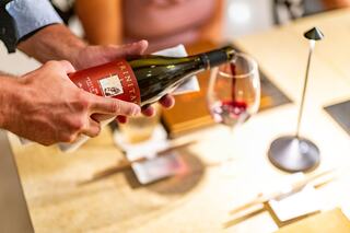 A hand pours a bottle of red wine into a glass, with a dimly lit table setting and another person in the background.