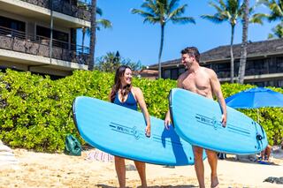 A cheerful couple carries surfboards along a sandy beach, surrounded by lush greenery and sunlight, ready for a fun day by the ocean.