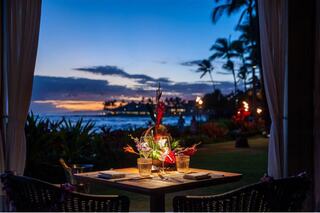 A tranquil dining setup by the ocean, adorned with flowers and candles, overlooking a colorful sunset and lush palm trees.