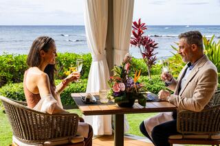 A couple enjoys a romantic toast at a seaside table, surrounded by lush greenery and vibrant tropical flowers, exuding elegance and joy.
